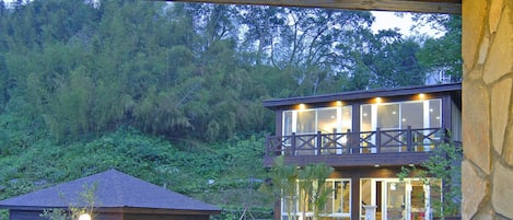 A wooden deck with two wicker stools and a glass table, overlooking a house with a stone wall and a balcony.