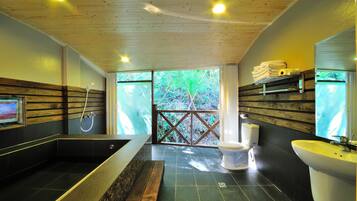 A modern bathroom with a bathtub, toilet, and sink, wooden ceiling, and a view of greenery outside.