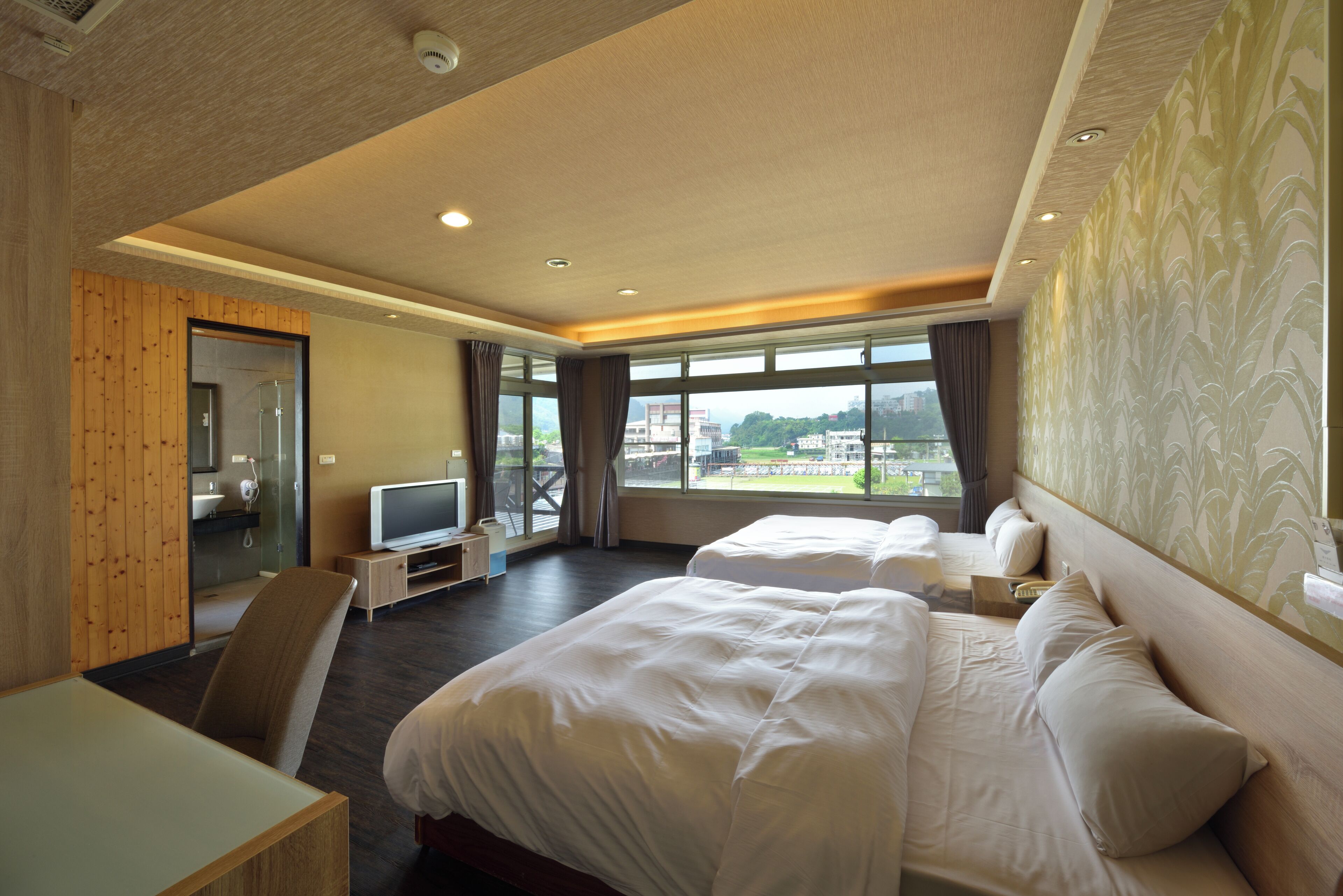 A hotel room with two beds, a TV, and a large window offering a city view.