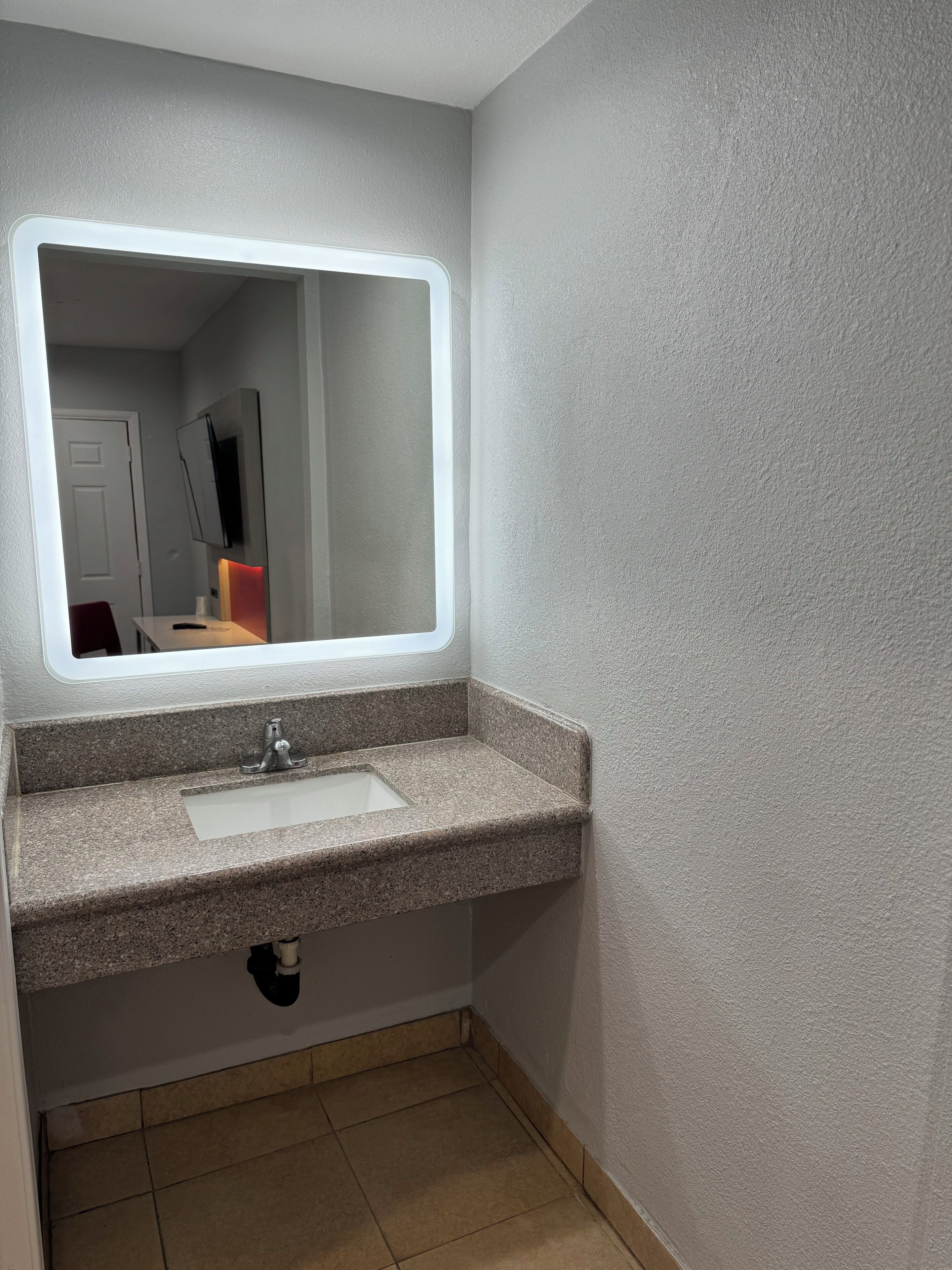 Une salle de bain avec un lavabo doté d’un plan de travail en granit, un miroir avec un éclairage intégré et une porte visible au fond.