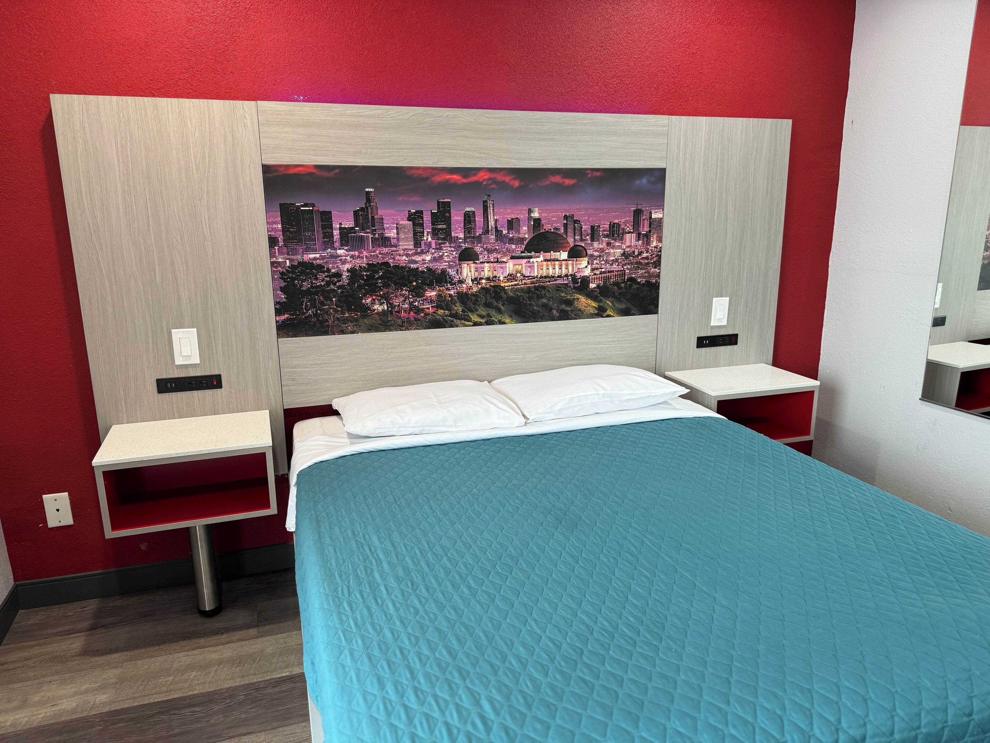 A hotel room with a bed, a nightstand, a wall-mounted TV, and a red wall.