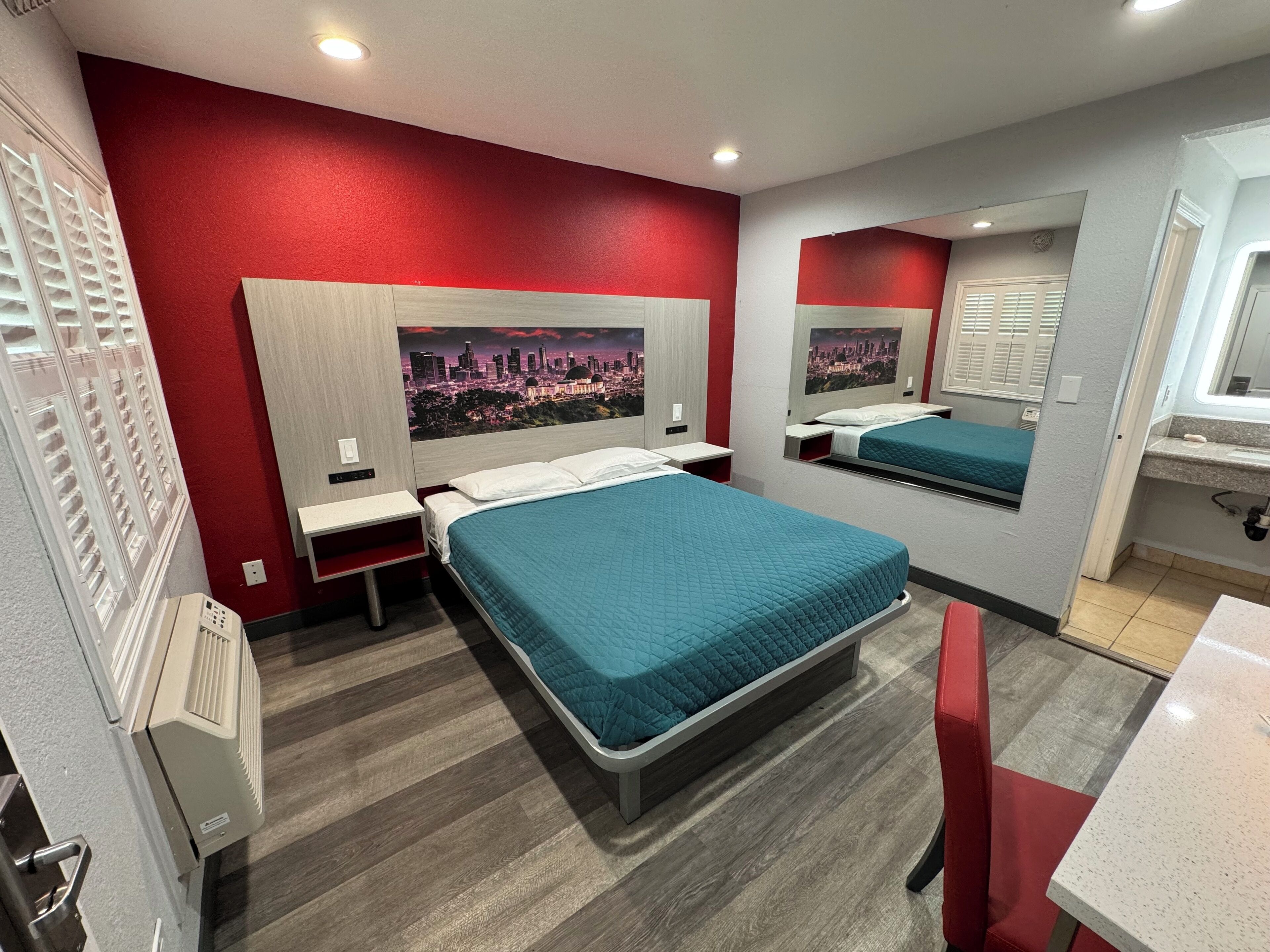 A hotel room with two beds, a red wall, a cityscape mural, and a bathroom with a sink and mirror.