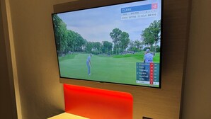 Uma televisão fixada na parede exibindo um campo de golfe, com uma poltrona vermelha estofada e um controle remoto sobre uma mesa abaixo.