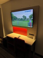Un televisor montado en la pared mostrando un campo de golf, con una butaca tapizada en rojo y un control remoto sobre una mesa debajo.