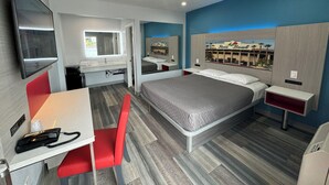 Quarto de hotel moderno com uma cama grande, uma mesa com telefone, uma cadeira, TV fixada na parede e um banheiro visível por uma porta aberta.