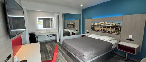 A modern hotel room with a large bed, a desk with a telephone, a chair, a TV mounted on the wall, and a bathroom visible through an open door.