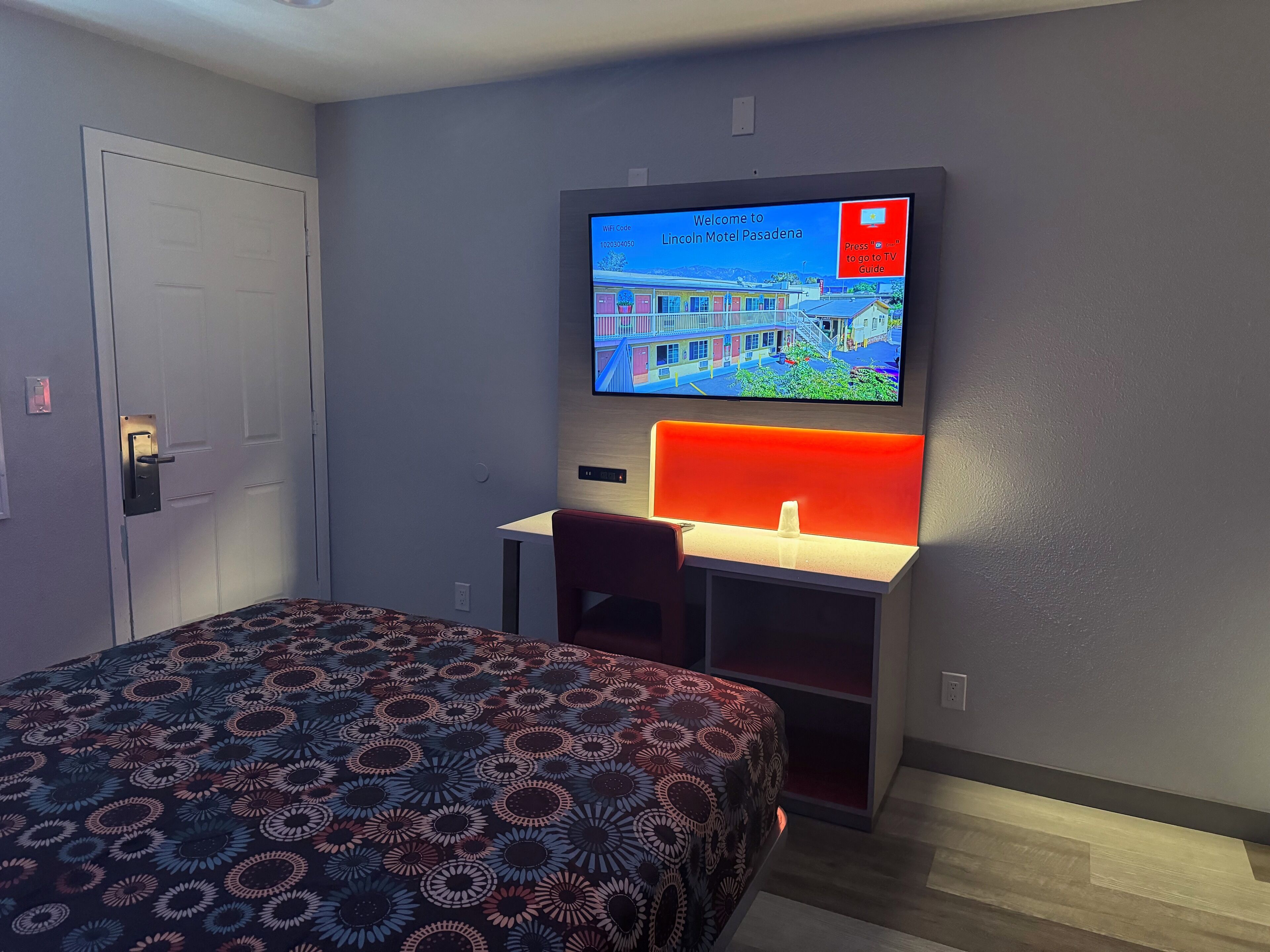 Quarto de hotel com cama, escrivaninha e painel vermelho iluminado, e televisão exibindo uma tela de boas-vindas.
