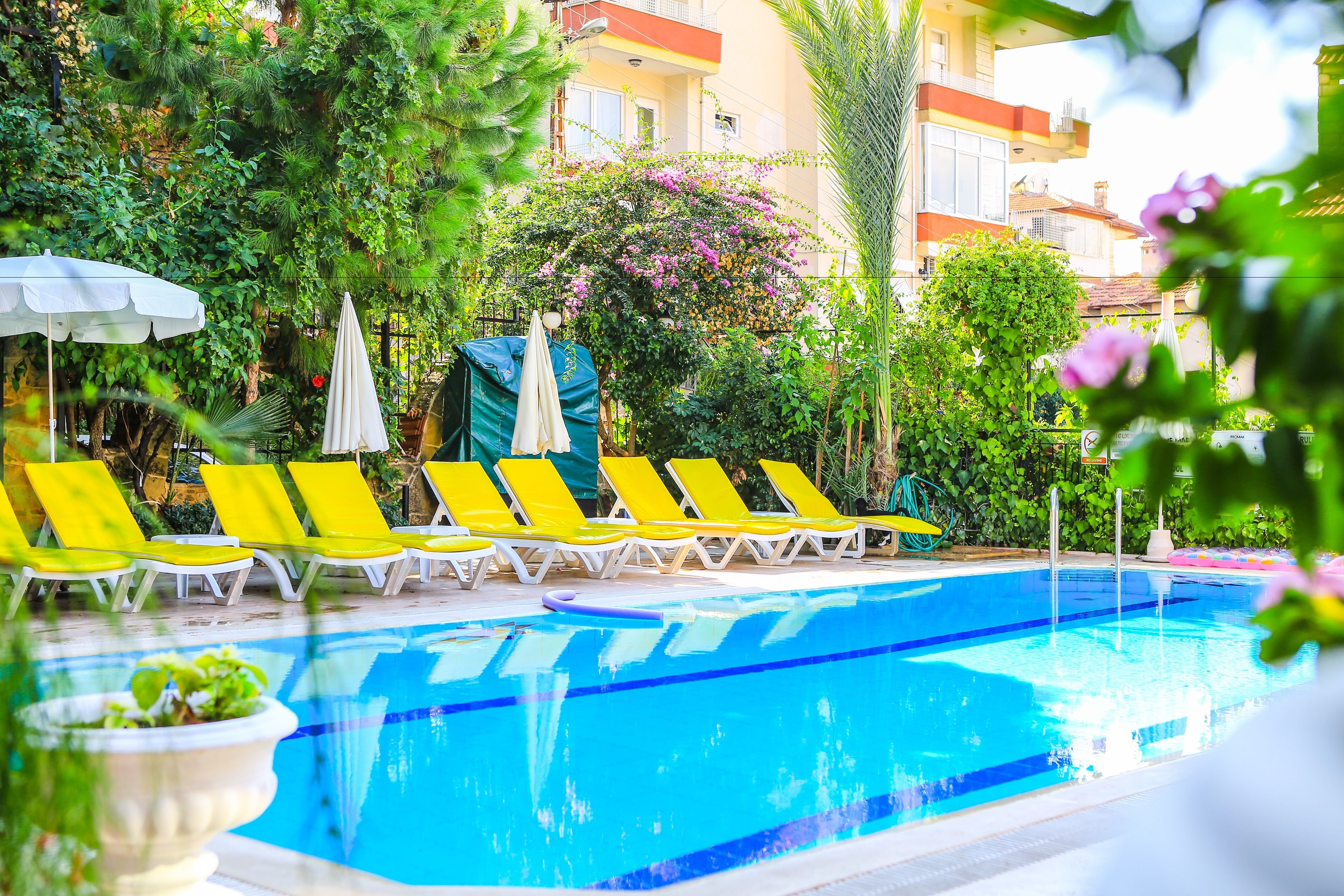 A swimming pool with yellow lounge chairs and umbrellas, surrounded by greenery and a building in the background.