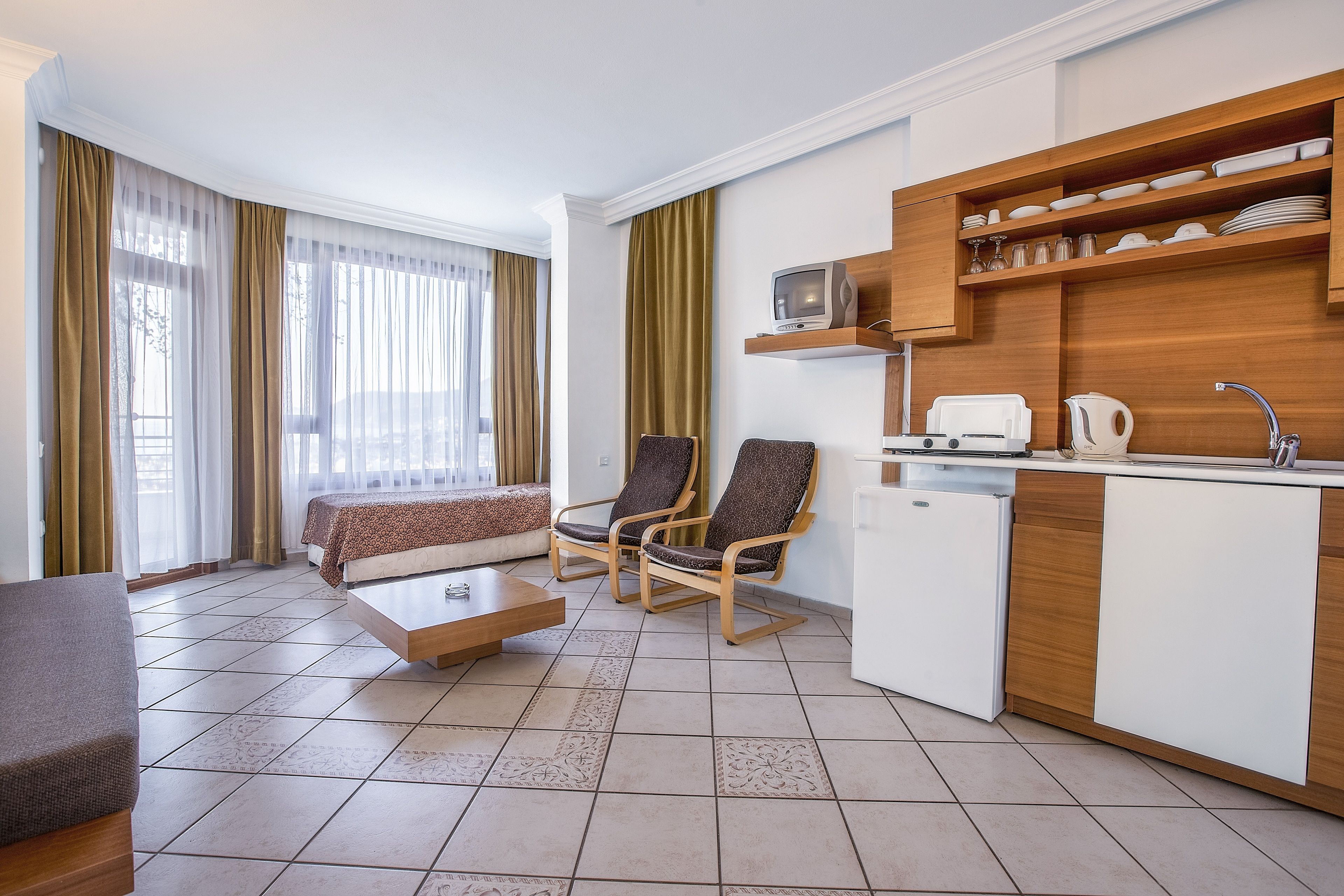 A modern hotel room with a sofa, two armchairs, a small coffee table, a TV, a mini-fridge, and a kitchen area with shelves and a sink.
