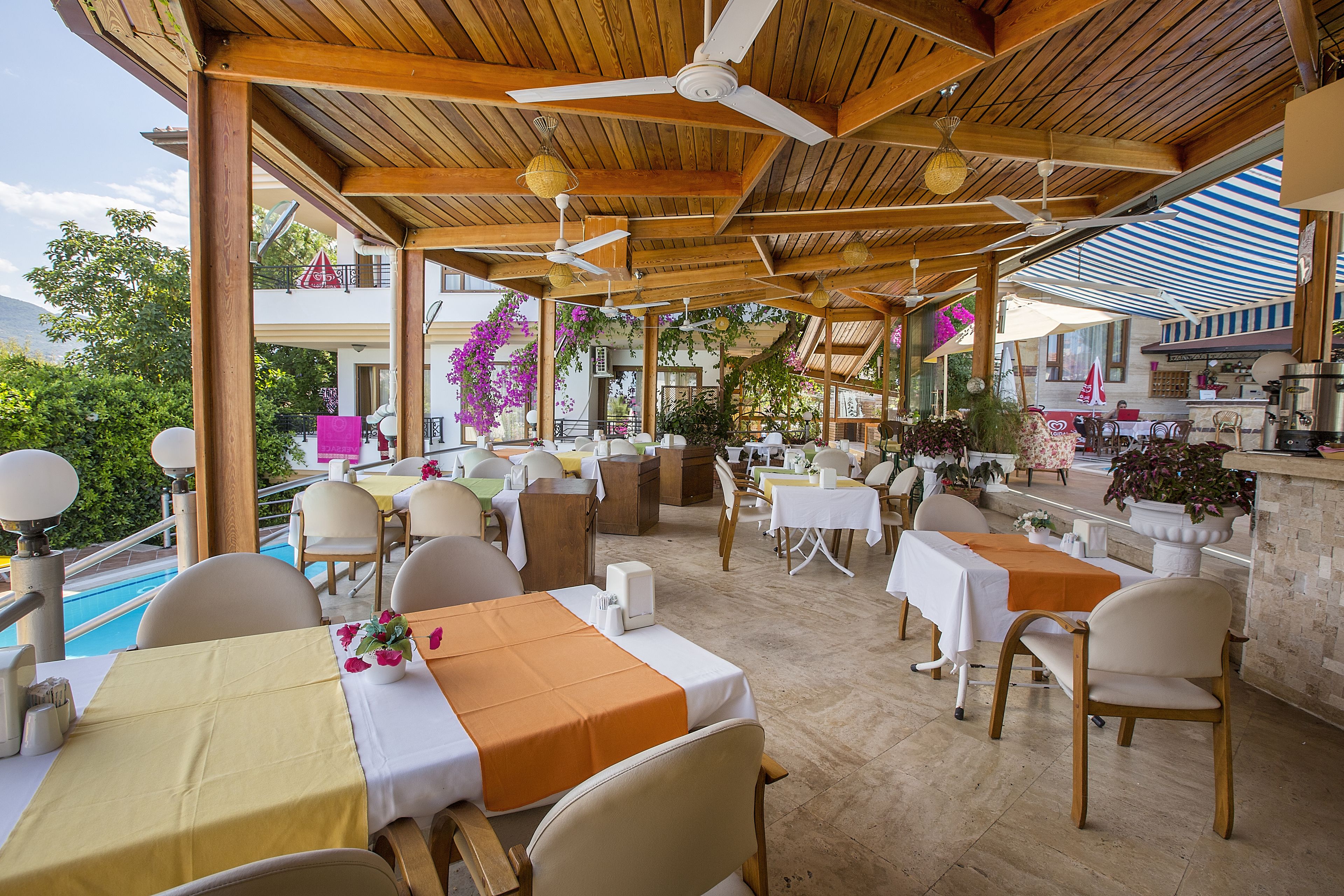 Un área de comedor al aire libre techada, con mesas y sillas, ventiladores de techo y plantas en macetas.