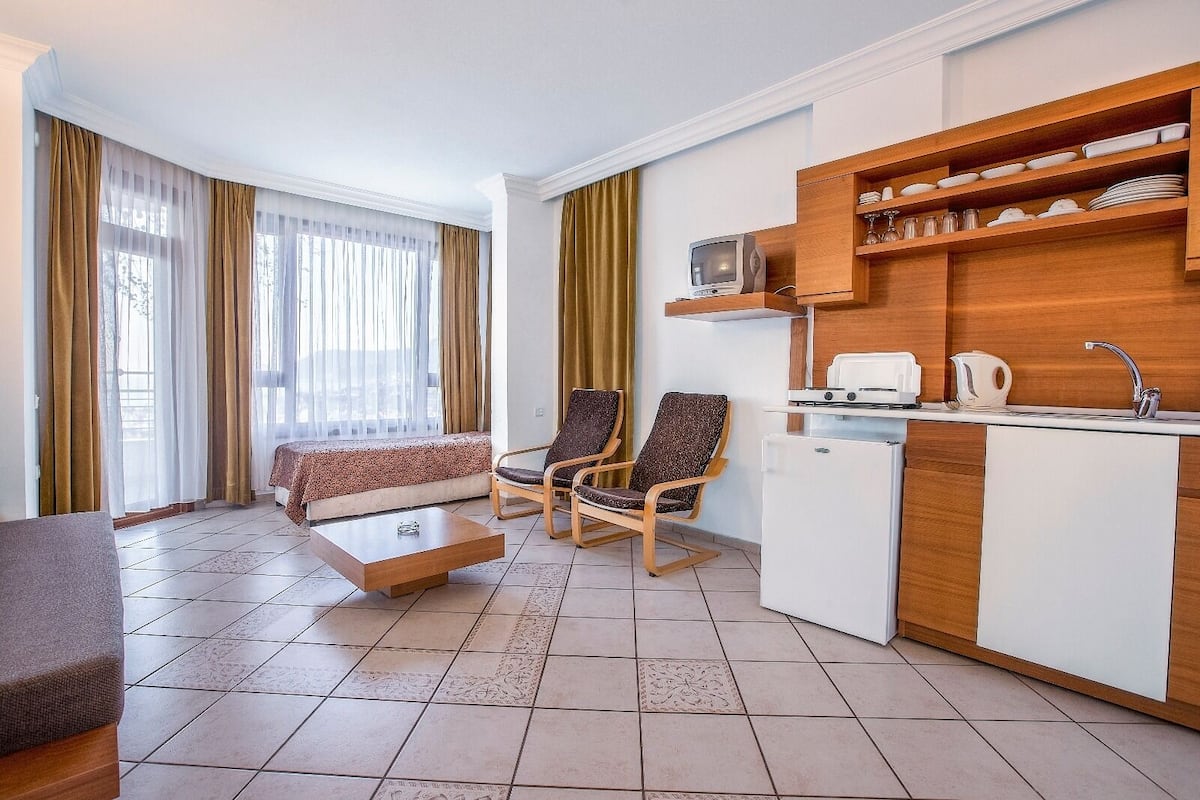 Un espacio habitable compacto con sofá, dos sillones, una mesita de centro, kitchenette con refrigerador, microondas, hervidor de agua y un lavamanos pequeño.