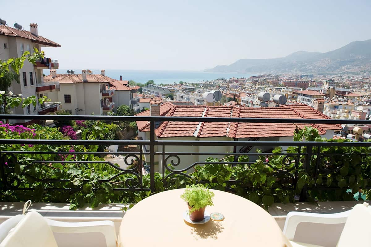 Un balcón con una mesa redonda, plantas en macetas y vistas de un paisaje urbano con edificios de tejados rojos y monta ñas al fondo.