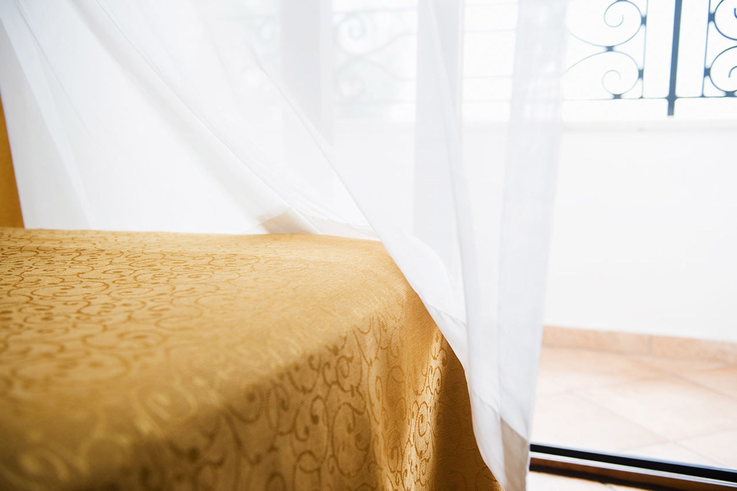 A bed with a patterned bedspread and sheer curtains partially drawn, revealing a balcony with a metal railing.