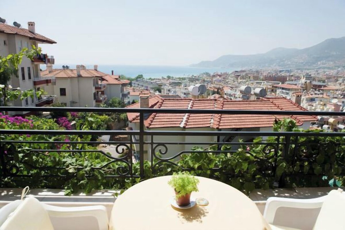 A balcony with a table and chairs, overlooking a cityscape with red-roofed buildings and a view of the sea.