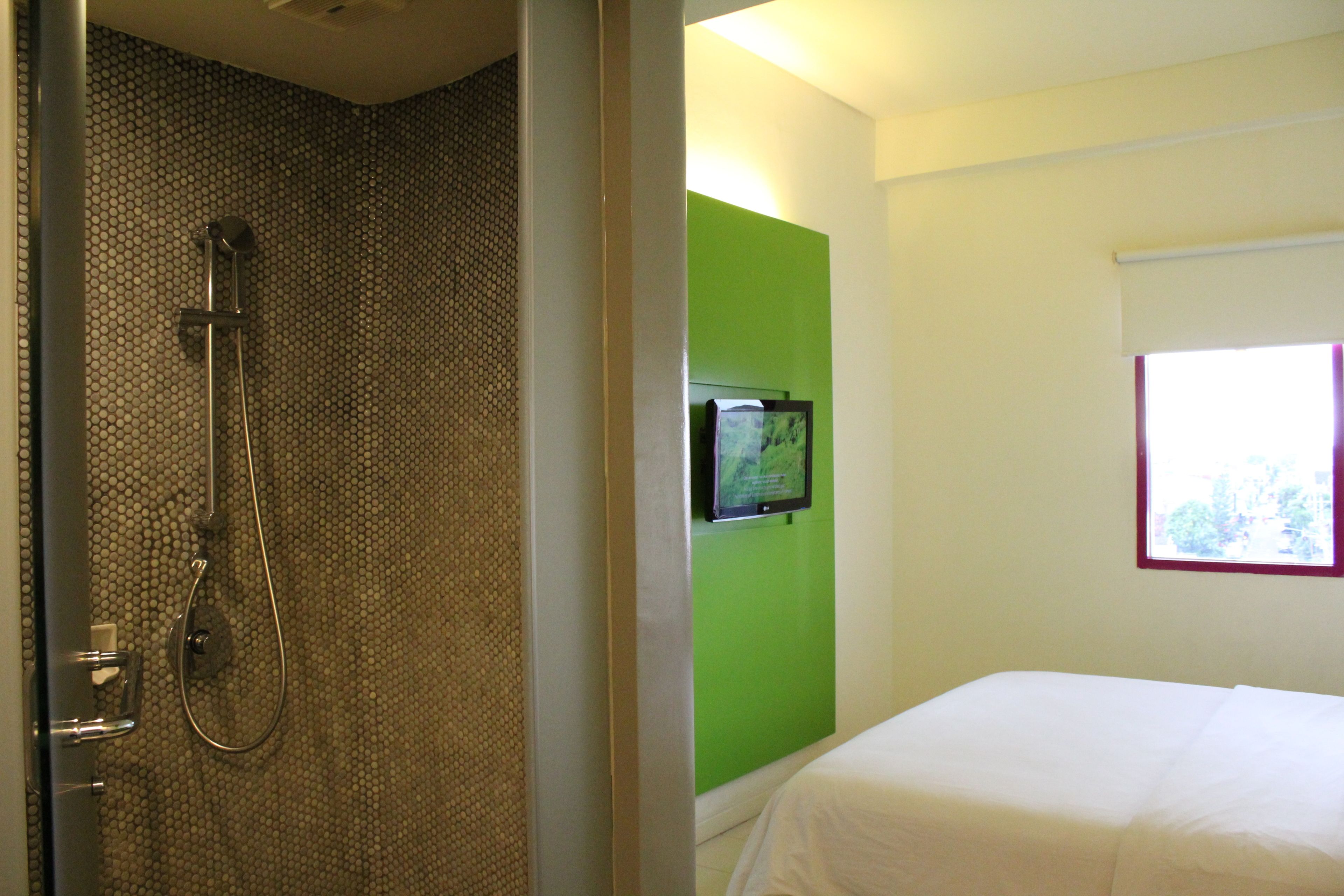 Une chambre d’hôtel avec une douche, un mur d’accent de couleur verte, un lit et une fenêtre avec des stores.