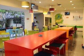Een helder verlichte ruimte met meerdere tafels en stoelen, een grote rode tafel op de voorgrond en een muurschildering op de wand.