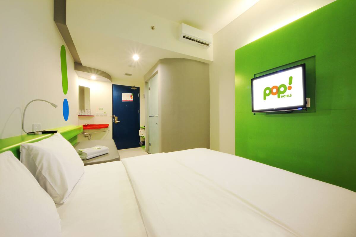 A hotel room with a bed, a green accent wall, a TV displaying 'pop! HOTELS' logo, and a bathroom with a blue door.