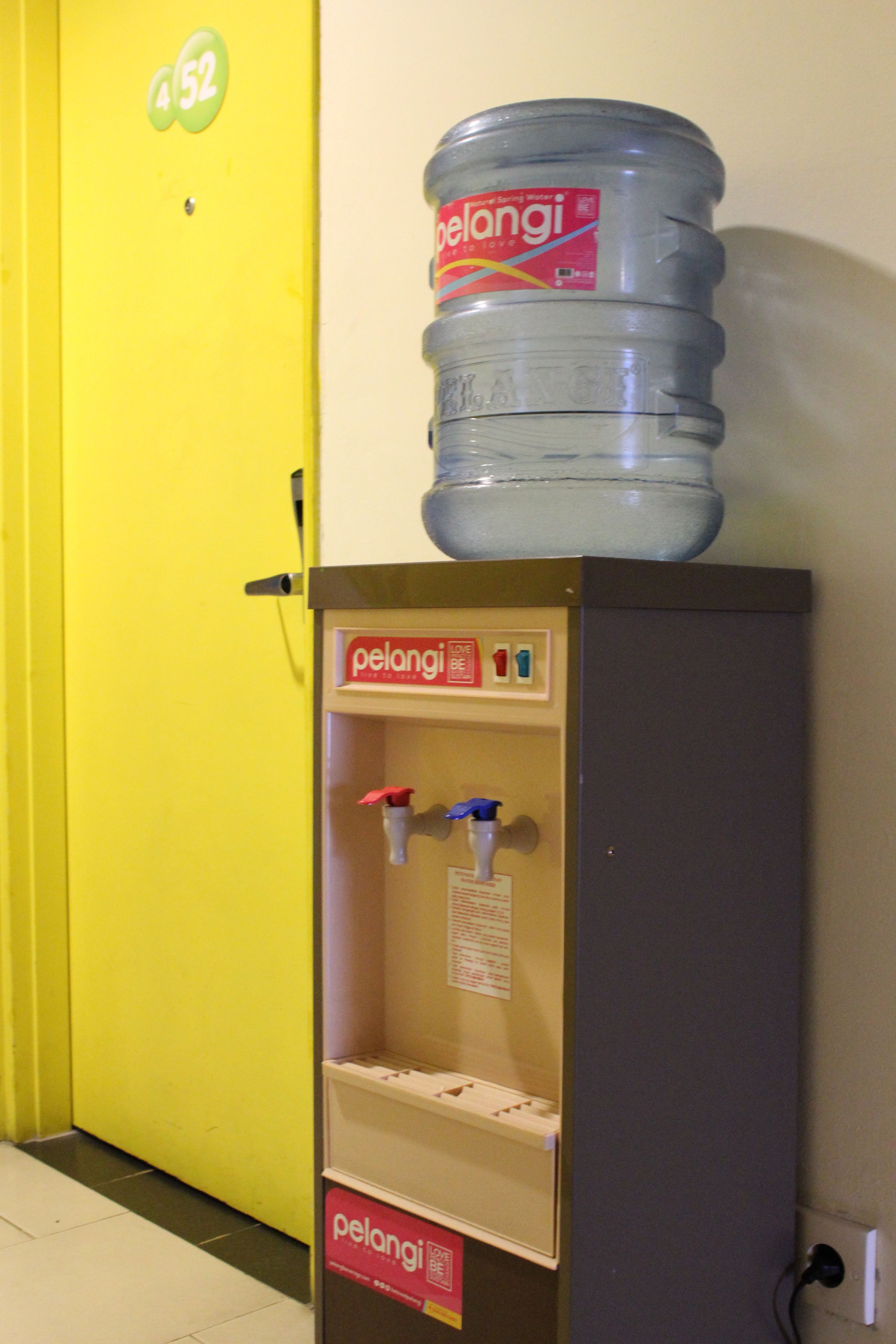 Un distributeur d’eau surmonté d’une grande cruche, situé dans un couloir avec une porte jaune et une pancarte indiquant le numéro de la pièce : 452.