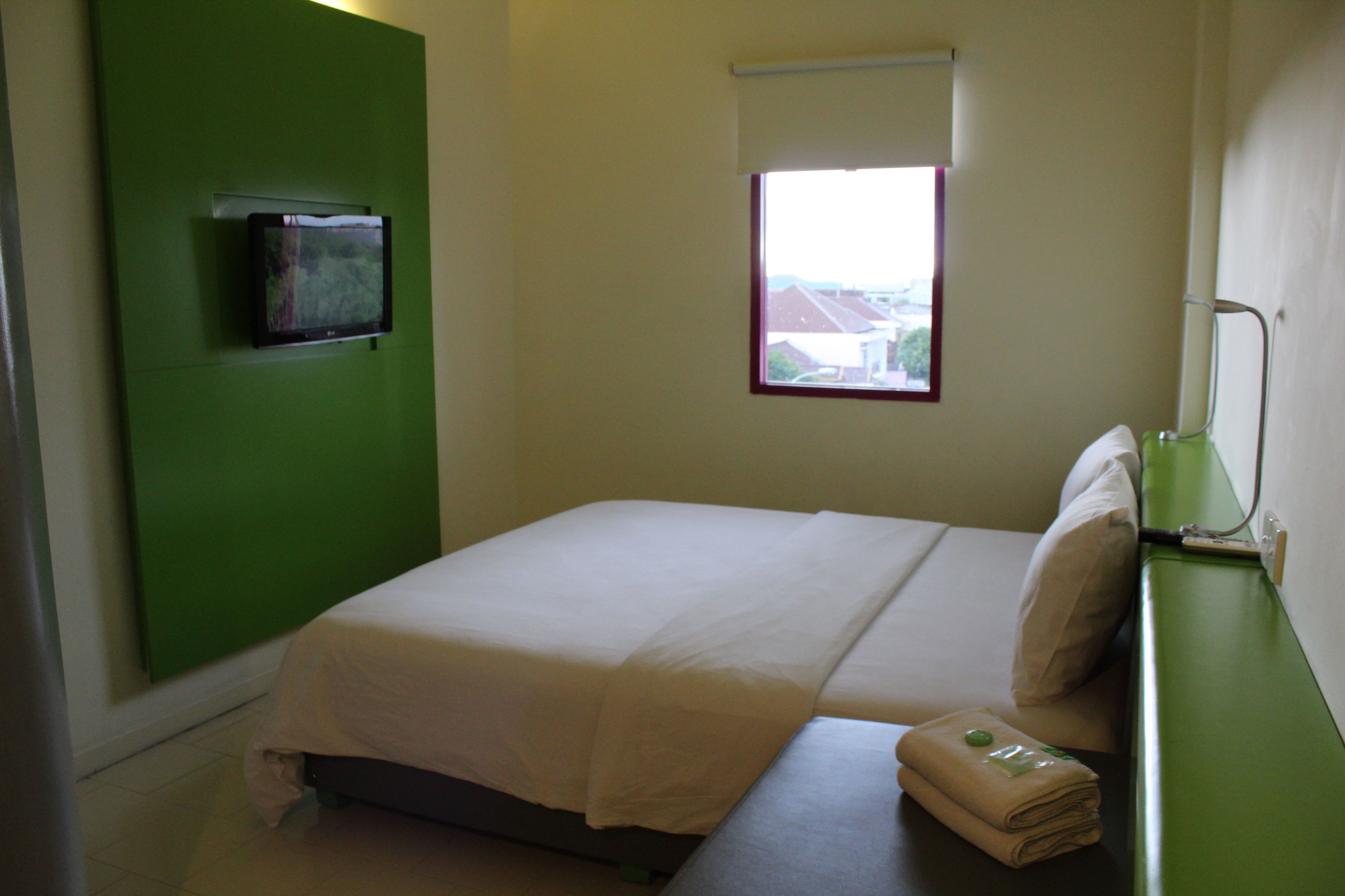 Un petit lit bien fait, avec un couvre-lit et des oreillers blancs, un mur vert orné d’un téléviseur à écran plat, et une fenêtre donnant sur les bâtiments environnants.