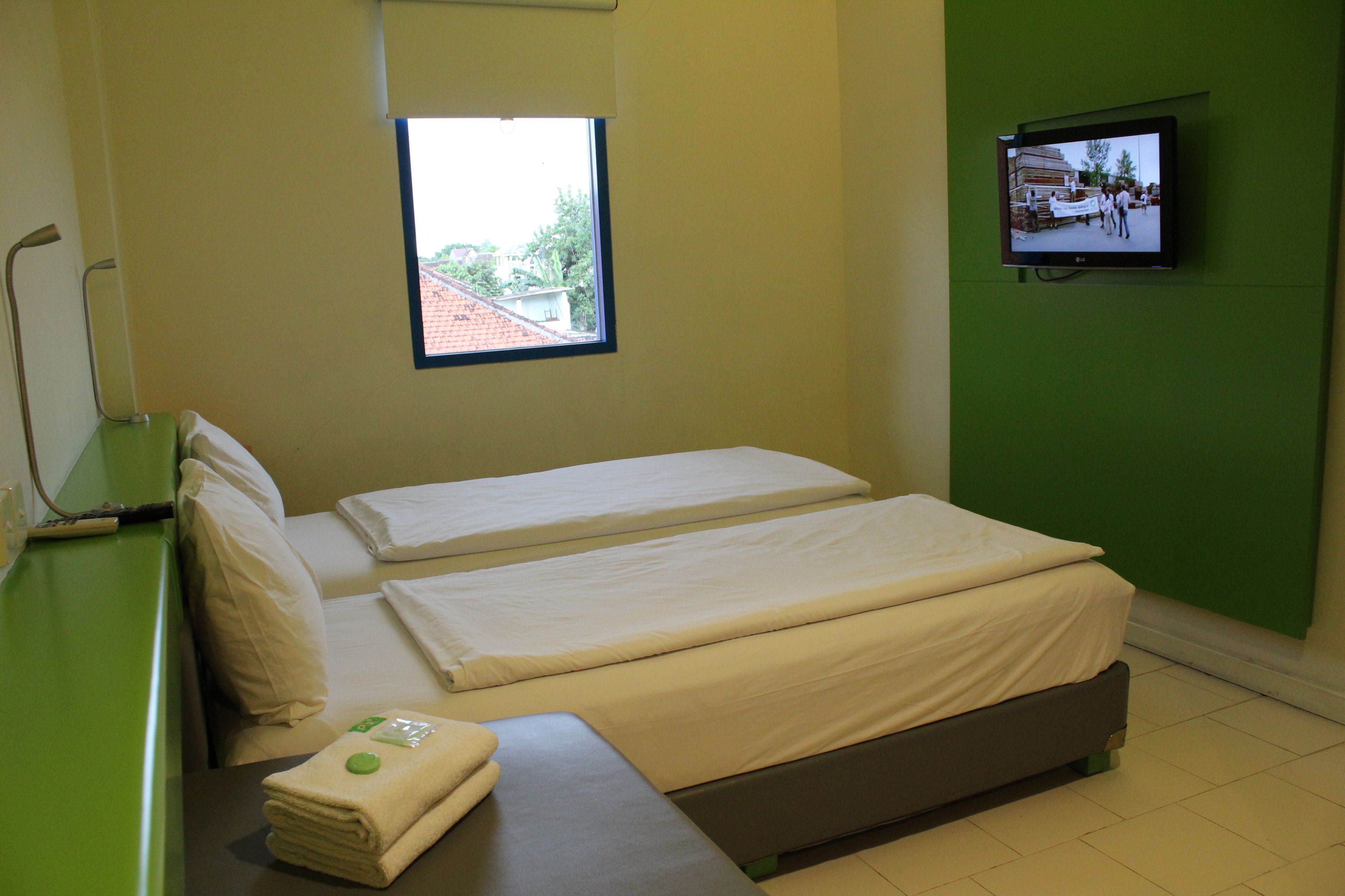 A hotel room with two single beds, a small table with a towel, a TV mounted on the wall, and a window providing a view of the outside.