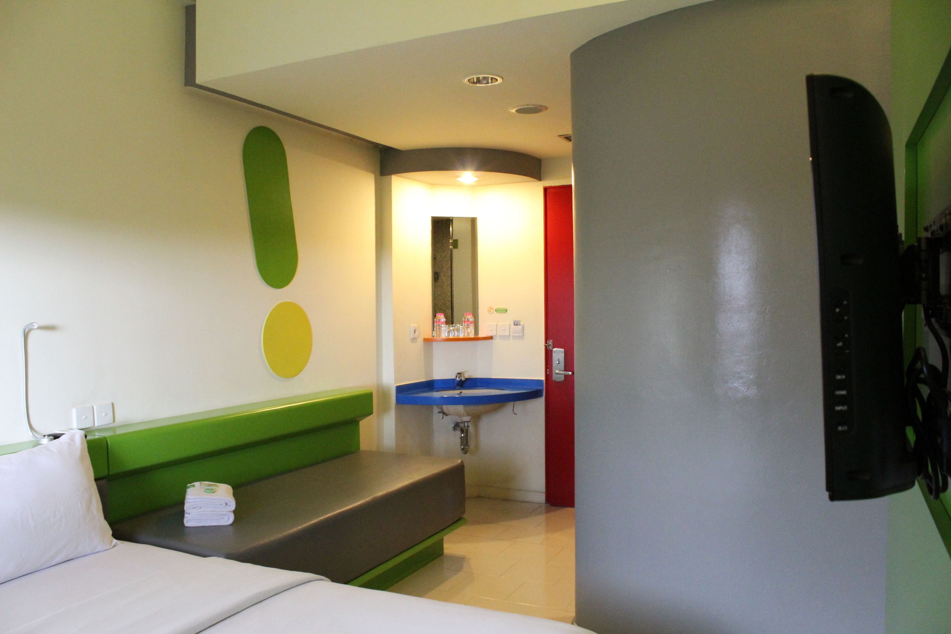 A hotel room with a bed, a green bench, a blue sink, and a red door.
