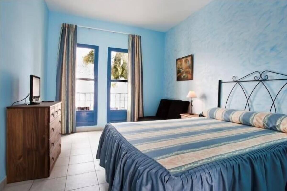 A bedroom with a bed, a desk, a dresser, and a window with curtains.