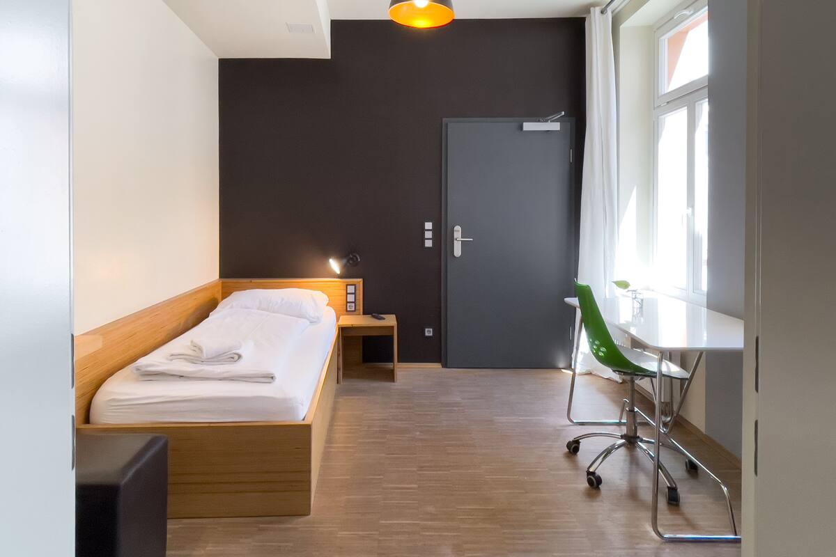 A modern hotel room with a wooden bed, a desk with a chair, a desk lamp, and a window with curtains.