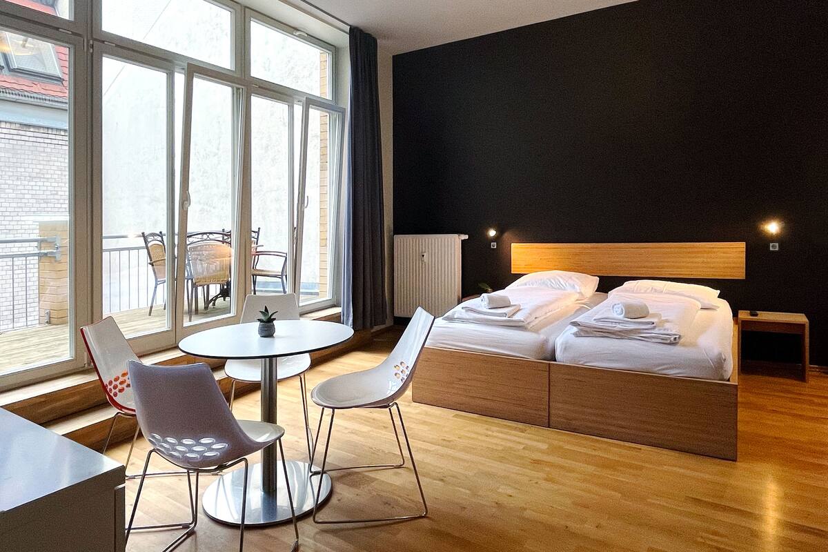 A modern hotel room with a large bed, a small table, two chairs, and a view of the outdoors through large windows.