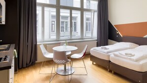 A hotel room with a bed, a small table with chairs, a window with curtains, and a view of buildings outside.