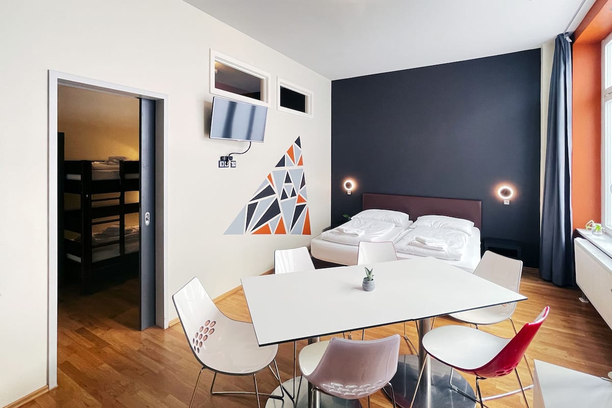 A modern hotel room with a bed, a dining table with chairs, a TV, and a geometric wall decoration.