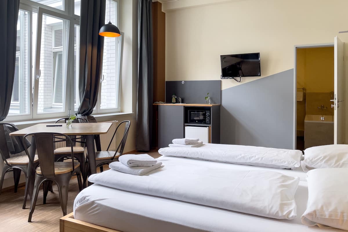 A hotel room with a bed, a desk with a chair, a small kitchenette, and a TV on the wall.