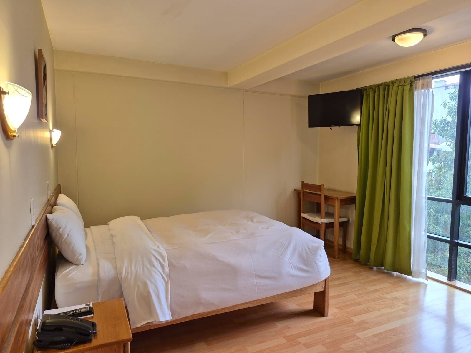 Ett hotellrum med en säng i trä, ett skrivbord, en stol, en platt-TV och ett fönster med gröna gardiner.