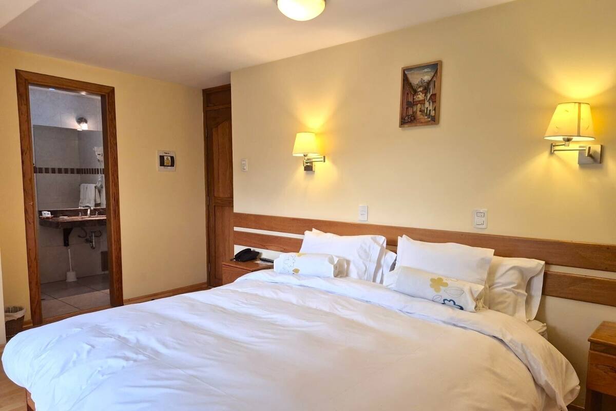 A hotel room with a large bed, two bedside lamps, a painting on the wall, and a bathroom visible through an open door.