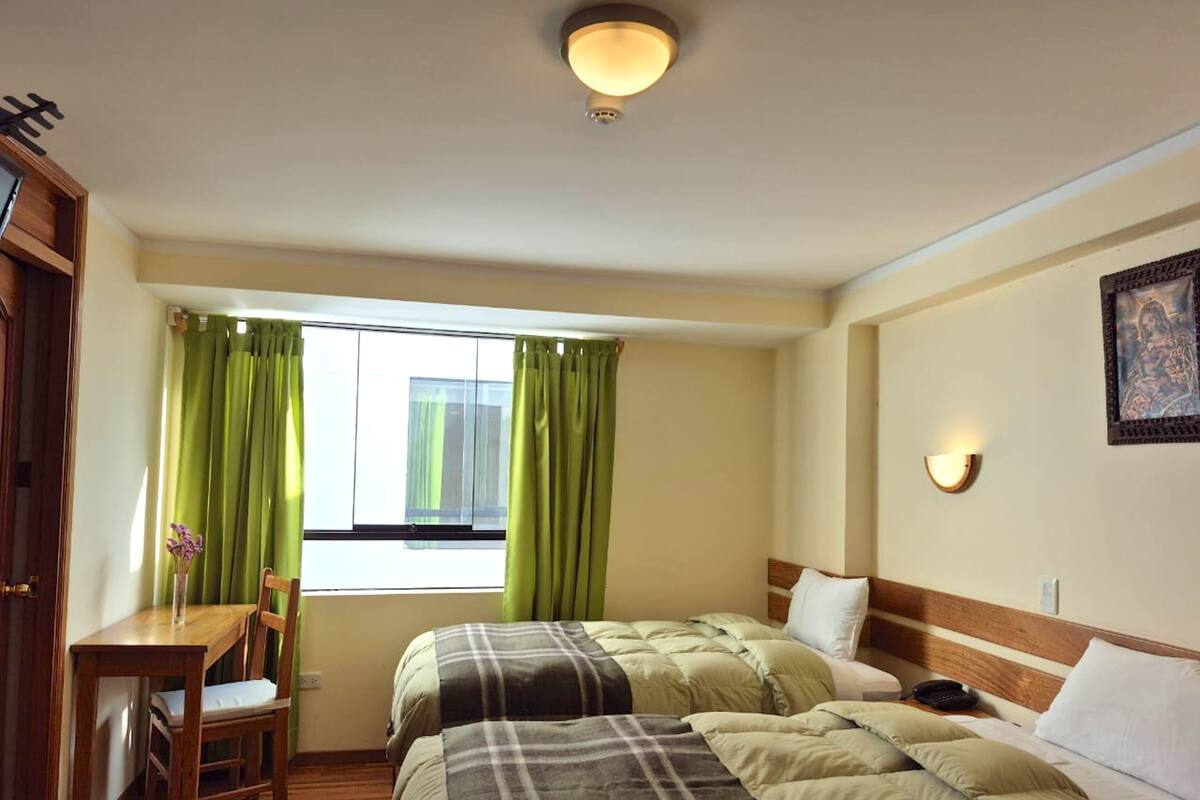 A hotel room with two beds, a desk, a chair, a window with green curtains, a wall-mounted light, and a framed picture.