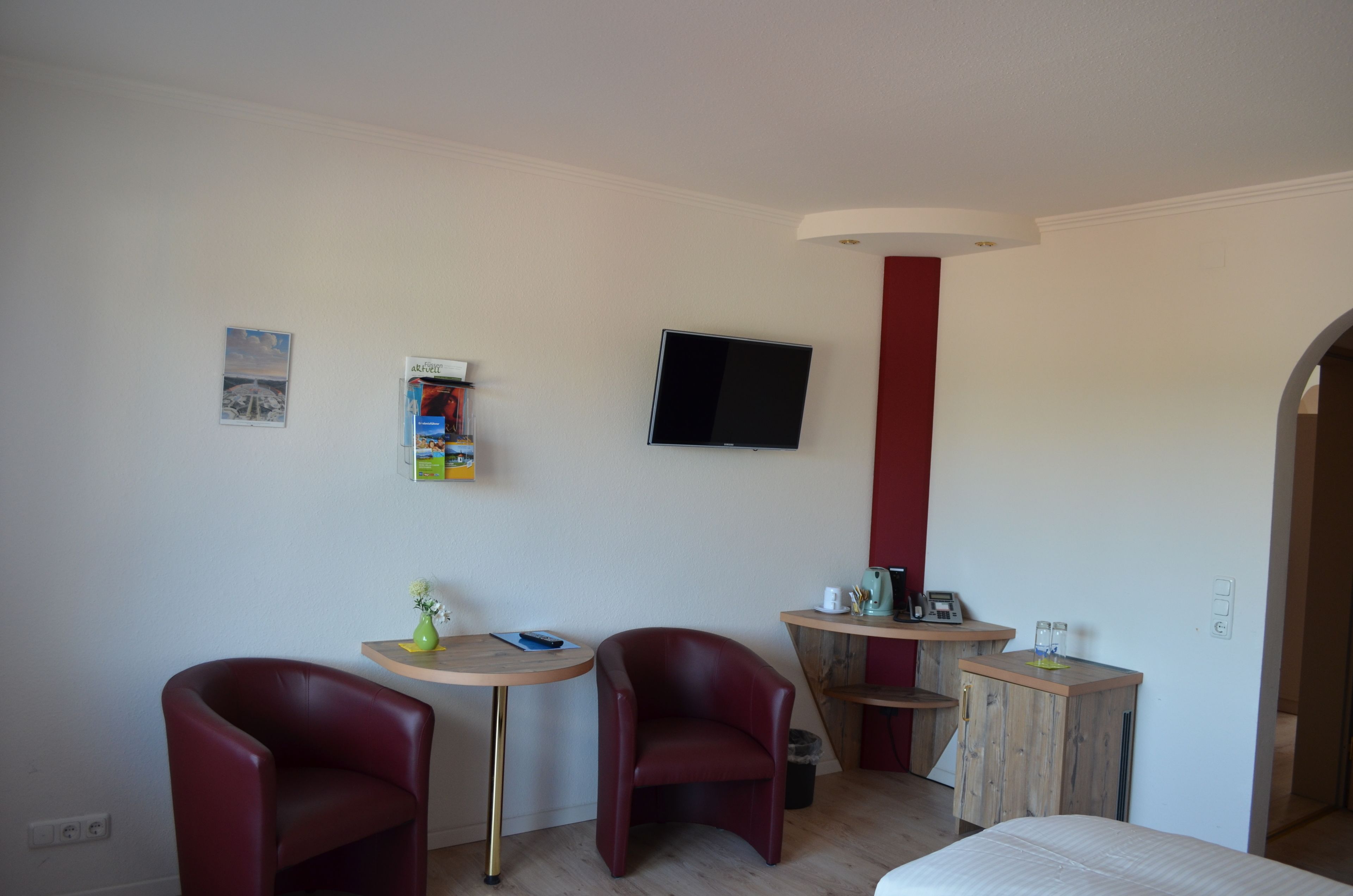A room with two red chairs, a small round table, a TV mounted on the wall, and a wooden cabinet.