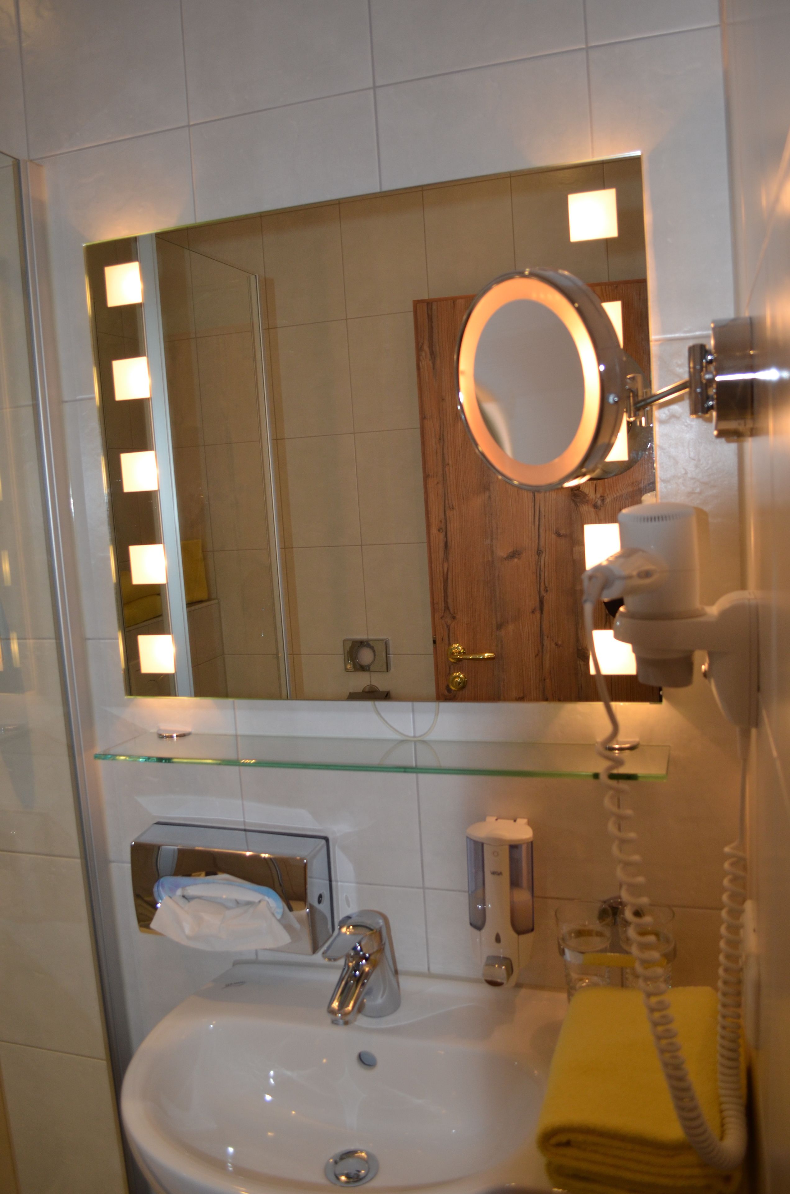Une salle de bain avec une étagère en verre, un lavabo, un miroir avec éclairage intégré, un sèche-cheveux et un porte-serviettes.