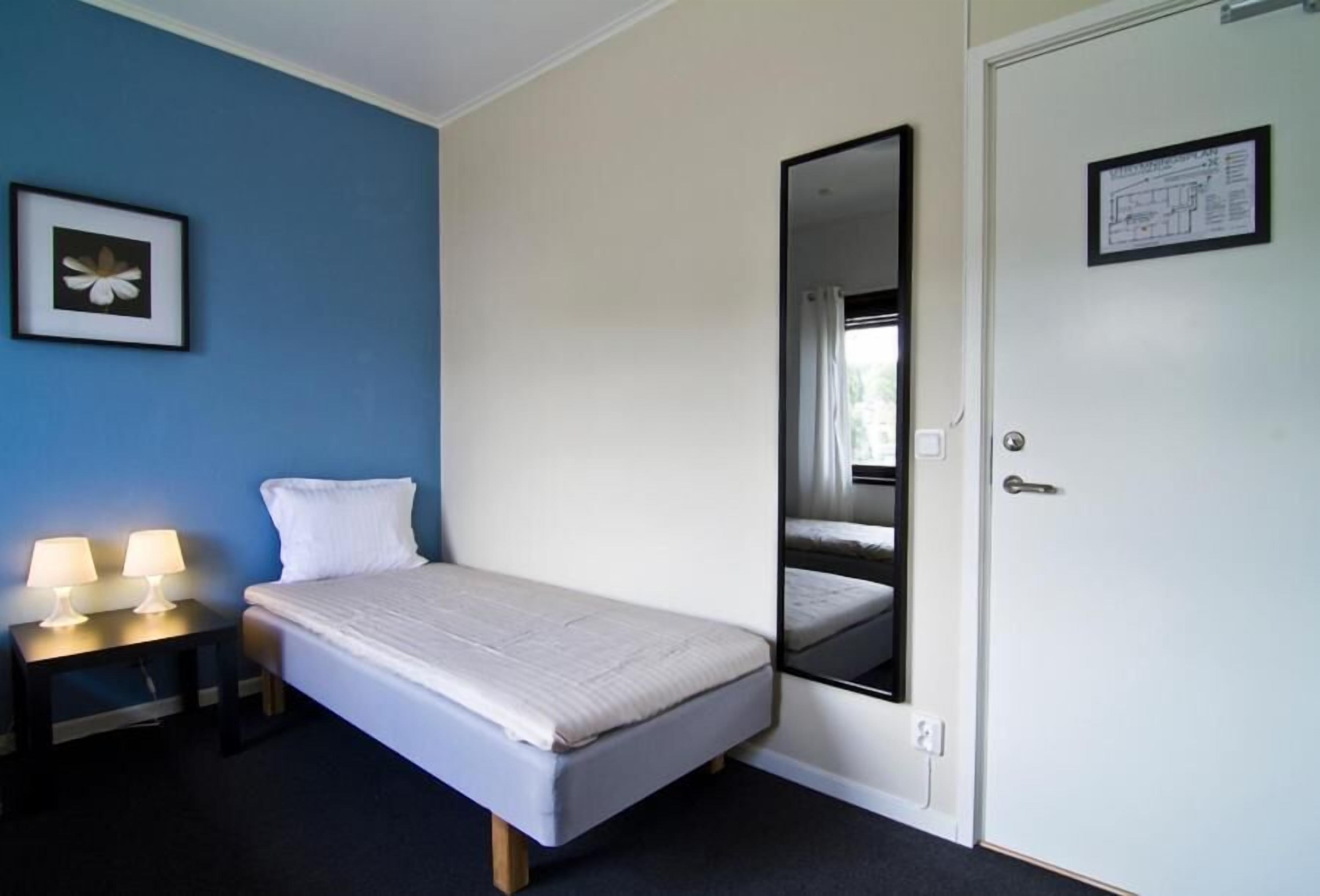 A single bed with a white mattress and pillows, a bedside table with two lamps, a mirror on the wall, and a door with a sign.