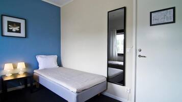A single bed with a white mattress and pillows, a bedside table with two lamps, a mirror on the wall, and a door with a sign.