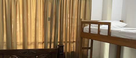 A room with two beds, one with a wooden headboard and the other with a bunk bed. There is a window with curtains, and a small table with a folded white cloth.
