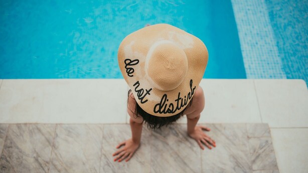 Una persona sentada sobre un suelo de baldosas cerca de una piscina, usando un sombrero de paja.