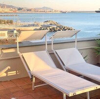 A balcony with two white sun loungers, a potted plant, and a view of the sea and distant cityscape.