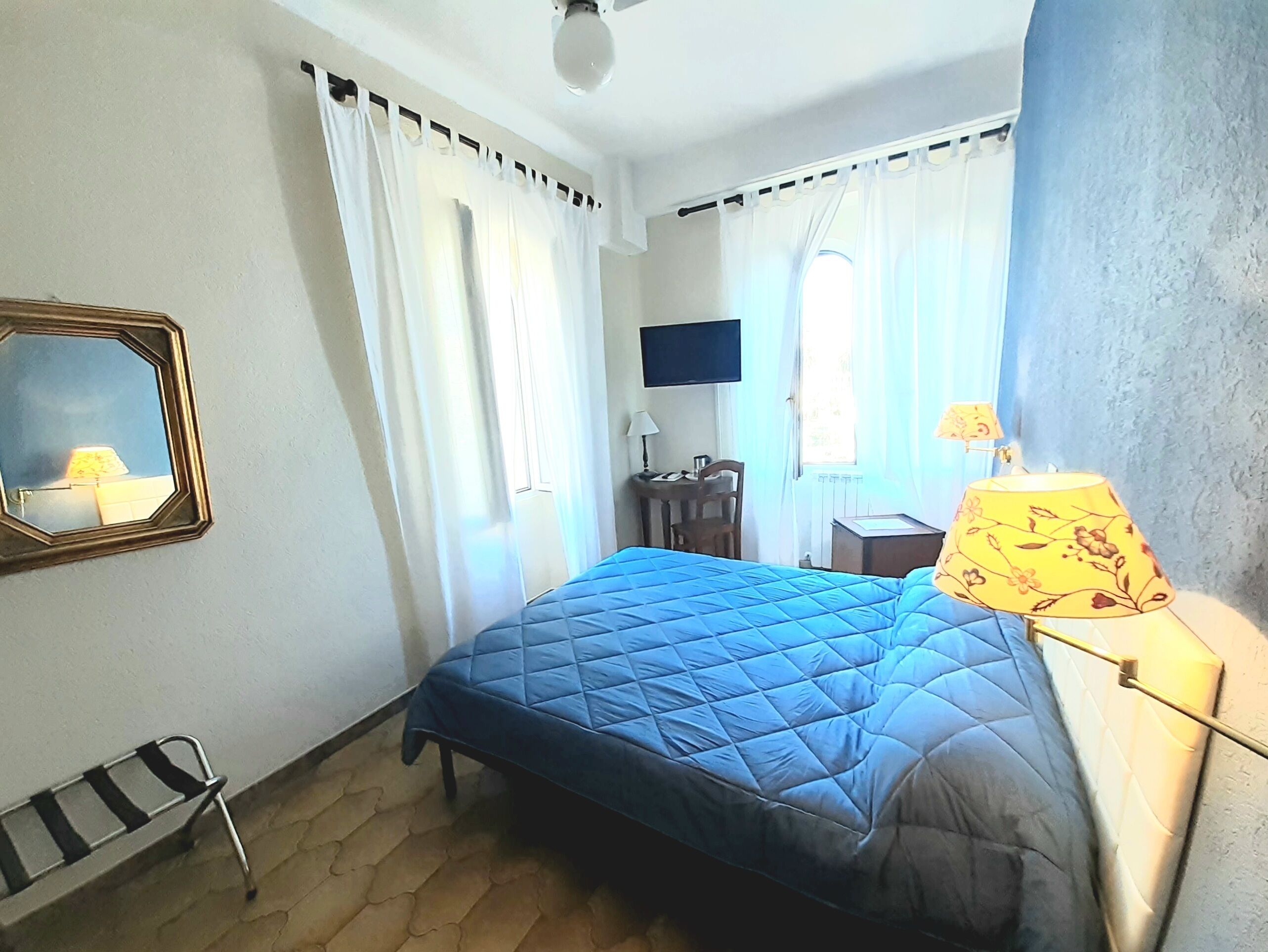 Un dormitorio con cama, una silla, un escritorio, una lámpara, televisión y ventana con cortinas.