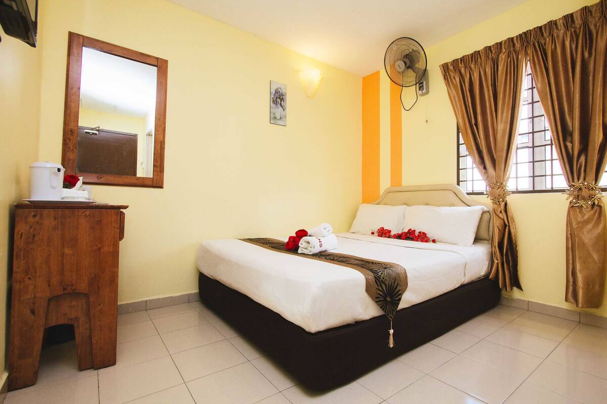 Een hotelkamer met een bed, een houten krukje, een spiegel, een ventilator en gordijnen.