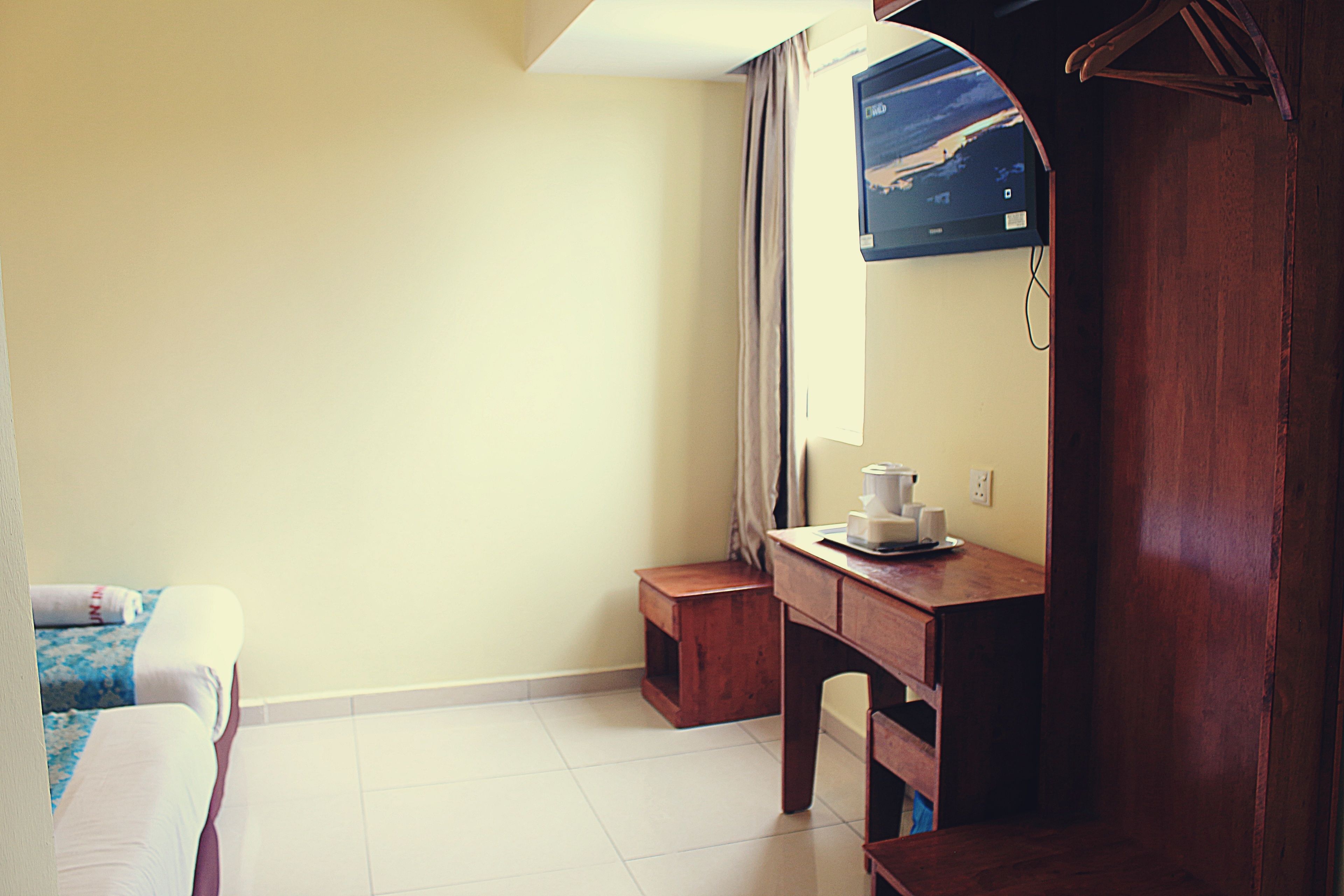 A hotel room with a bed, a wooden desk, a small wooden stool, a wall-mounted TV, and a window with curtains.