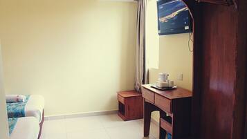 A hotel room with a bed, a wooden desk, a small wooden stool, a wall-mounted TV, and a window with curtains.