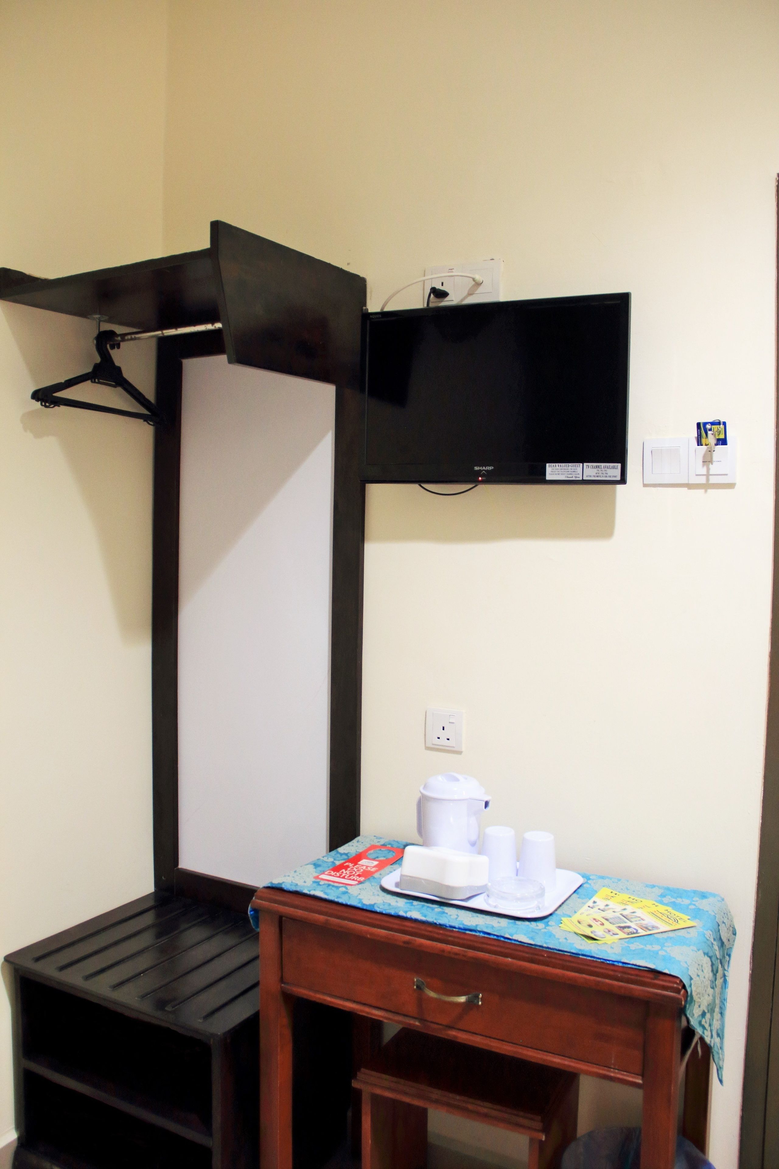 Een kleine kamer met een houten bureau, een zwarte kapstok, een flatscreen-tv en een stopcontact.