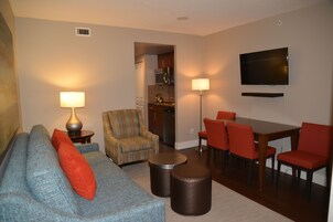 A living room with a sofa, armchair, coffee table, dining table with chairs, a flat-screen TV, and a kitchen area.