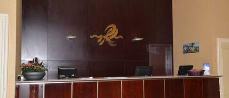 A reception desk with a dark wood finish, a large decorative letter 'R', and a potted plant on the left.