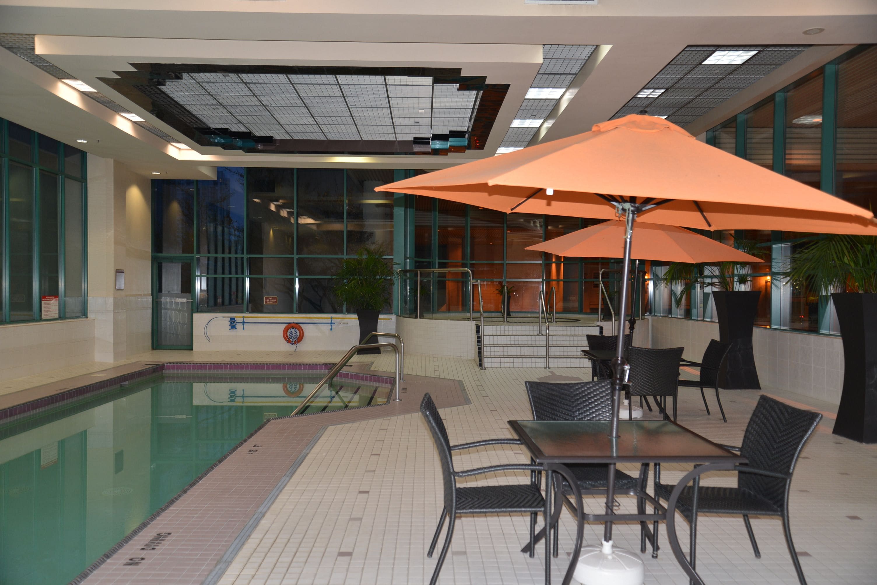 Un espace piscine intérieur doté d’une table et de chaises, entouré de murs en verre et surmonté d’un plafond agrémenté de lucarnes.