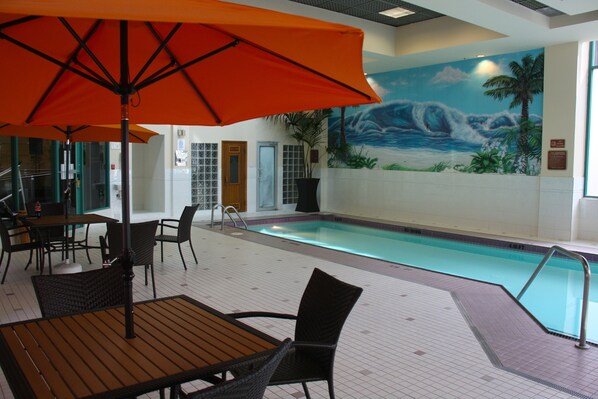 Uma área de piscina interna com um mural de uma cena de praia, um espaço de estar à beira da piscina com mesas e cadeiras, e um grande guarda-sol laranja oferecendo sombra.